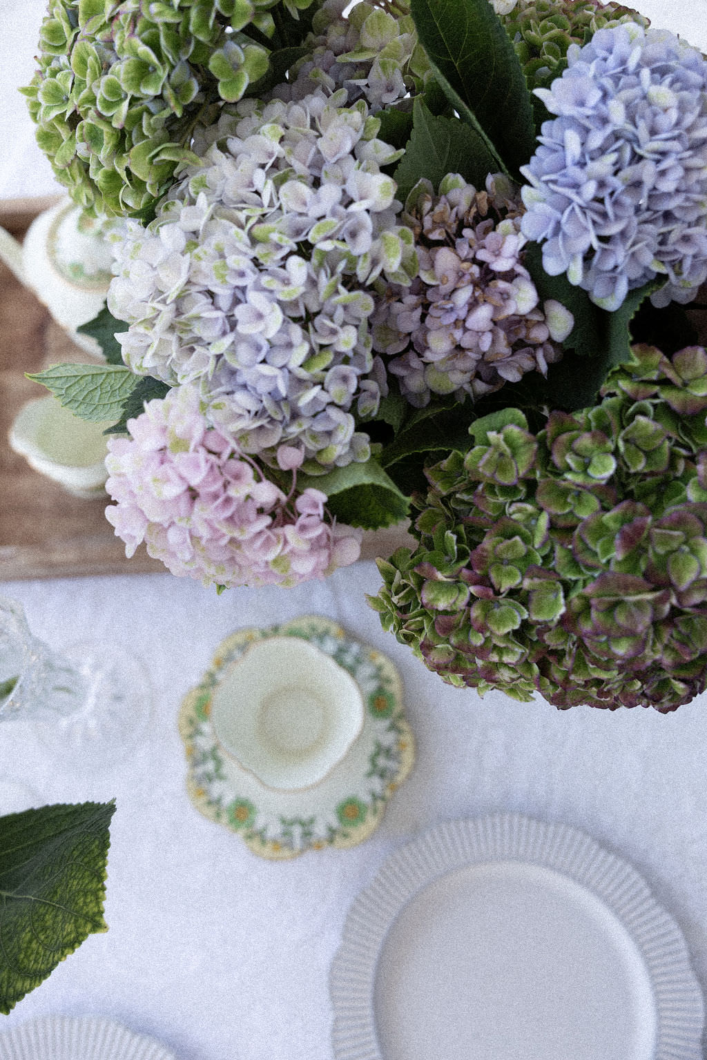 Beautiful purple and green hydrangea bouquet arranged beside elegant vintage teacup and saucer on white linen tablecloth"